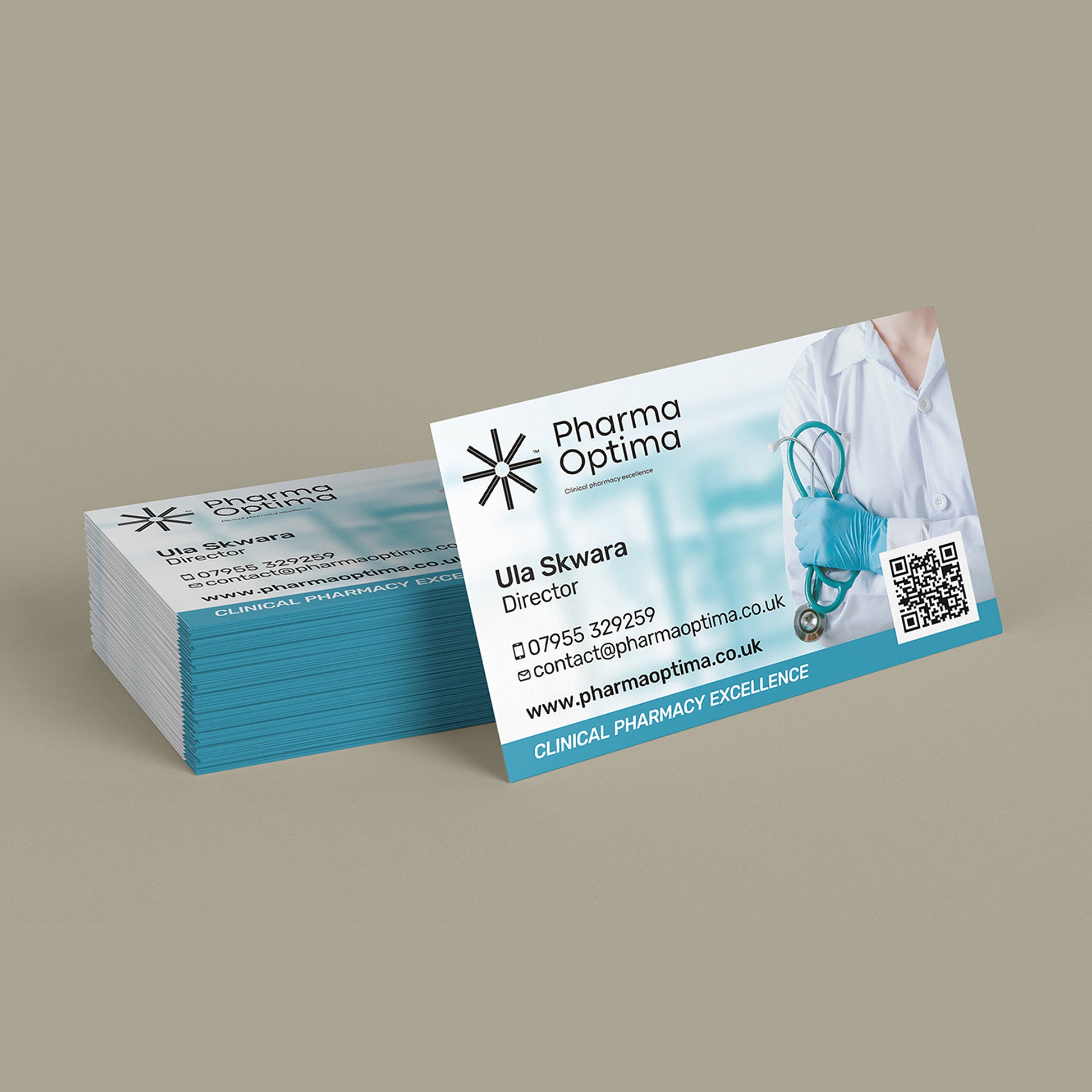 A stack of business cards for Pharma Optima Pharmacy, with one card standing upright in front. The card design is white and teal with text including 'Ula Skwara Director' and a stethoscope photo, photographed on a plain taupe background.