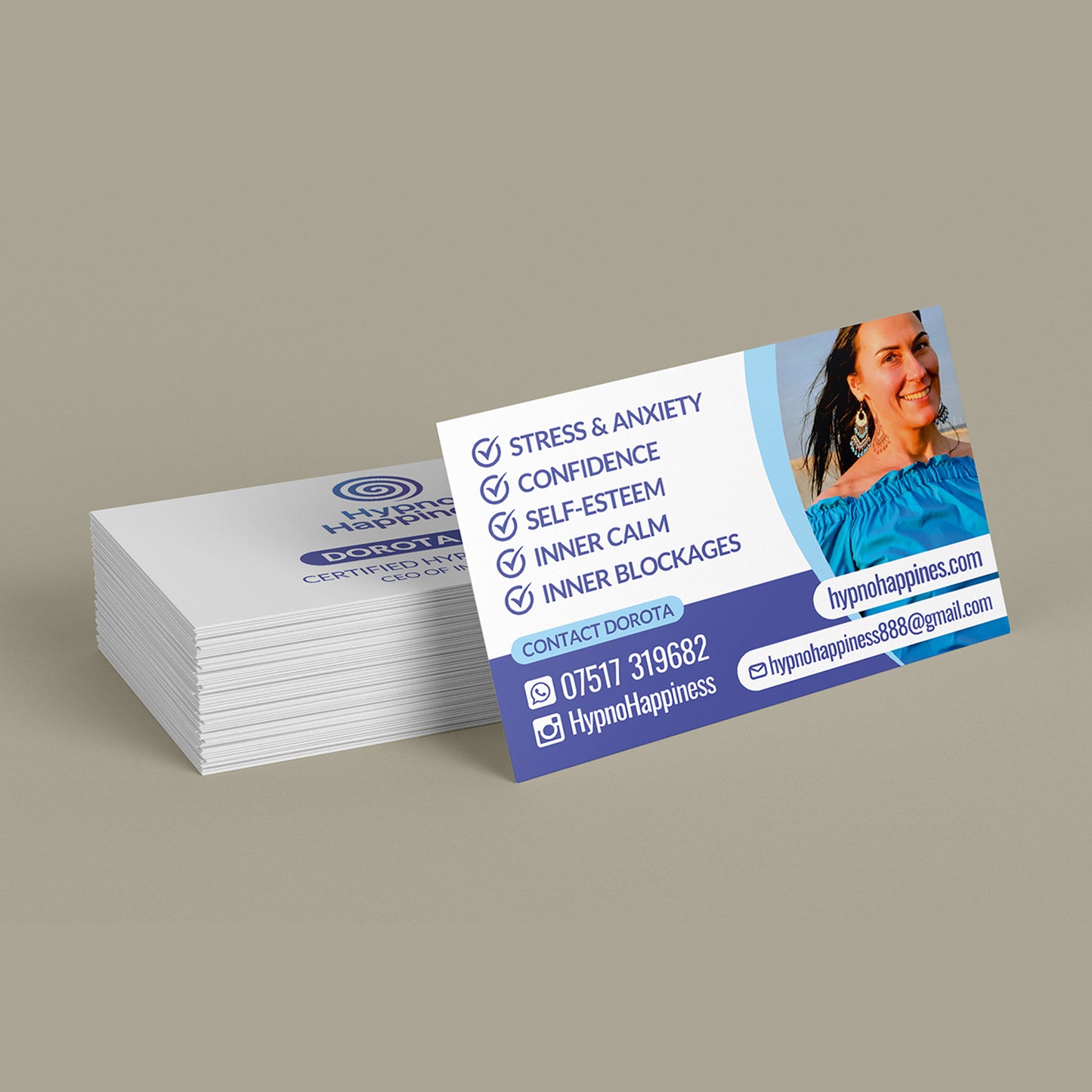 A stack of business cards for Hypno Happiness, with one card standing upright in front. The cards are white and blue with text listing benefits like stress and anxiety, confidence, self-esteem, and inner blockages, alongside a photo of a smiling woman in a blue top.